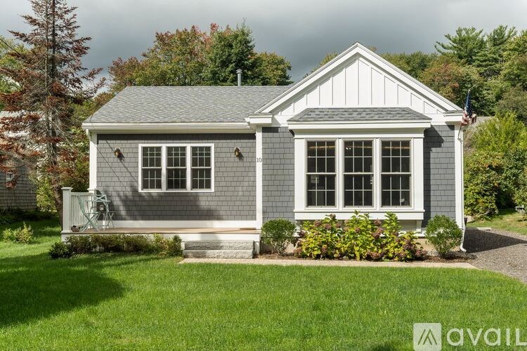 A small house with a grey roof and white trim is surrounded by greenery.
