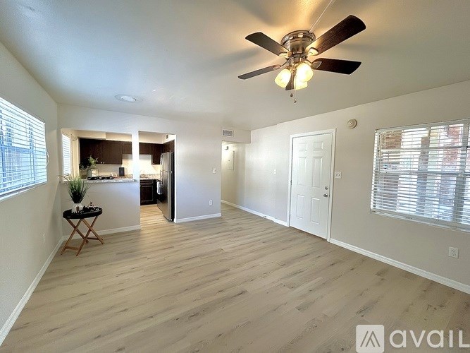 A spacious room with a ceiling fan and light, hardwood floors, and a view of a kitchen in the background.