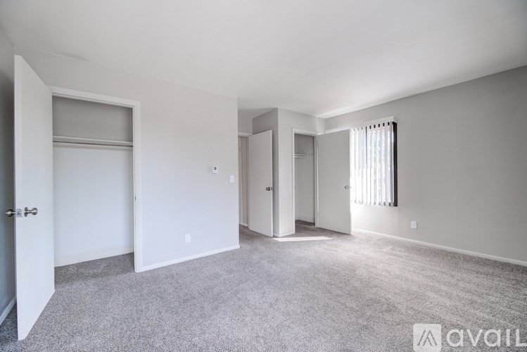 A spacious room with a carpeted floor and white walls, featuring a door, a window with blinds, and a wall-mounted light switch.