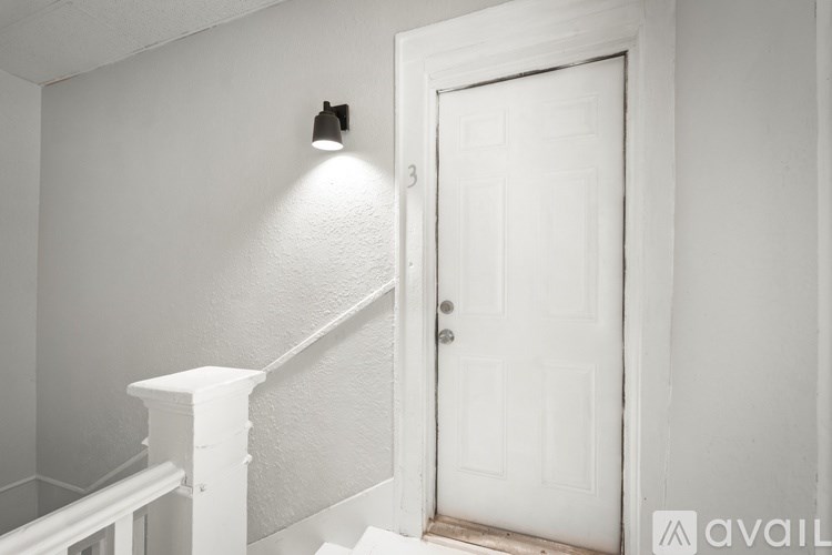 A white door is in a white room with a light on the wall.
