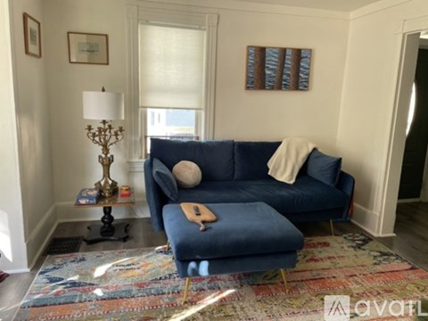 A blue couch with a white pillow and a tan blanket is in the middle of a room with a rug and a lamp.