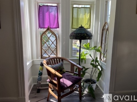 A wooden chair with a purple cushion sits in front of a window with a purple curtain.