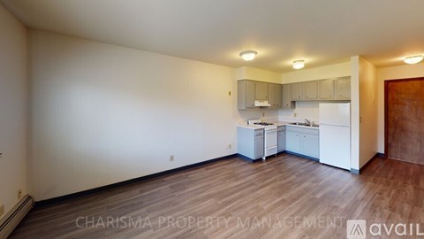 A well-lit, empty room with wooden flooring and a kitchenette in the background.