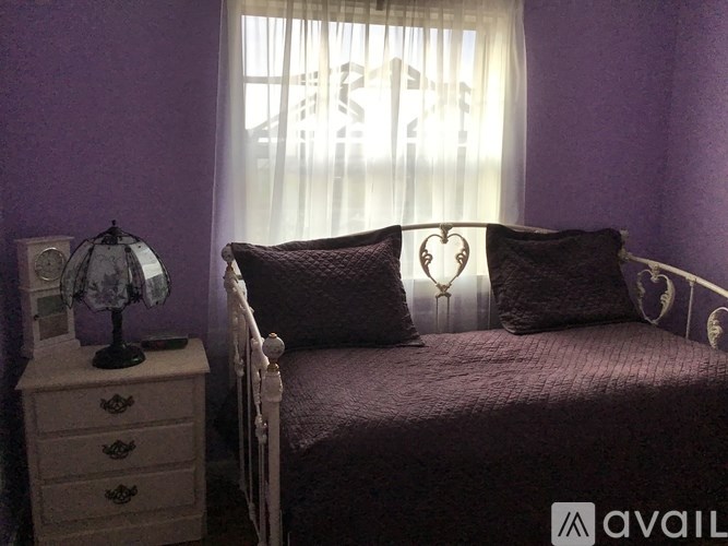 A bed with a purple blanket and two heart-shaped pillows sits next to a nightstand with a lamp on it.