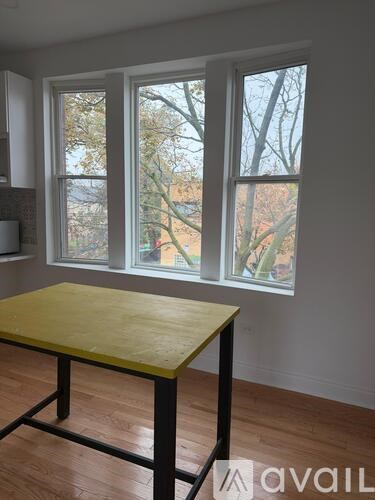 A table with a yellow top is in front of a window with a view of trees and a building outside.