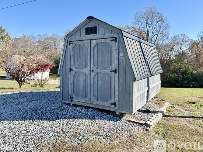 A grey barn with a metal roof is situated in a gravel area with a grassy field and trees in the background.