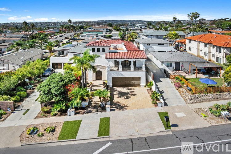 A large house with a red tile roof is surrounded by a well-manicured lawn and other houses in the background.