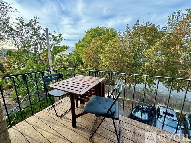 A wooden table and two chairs are set up on a balcony with a view of cars and trees.