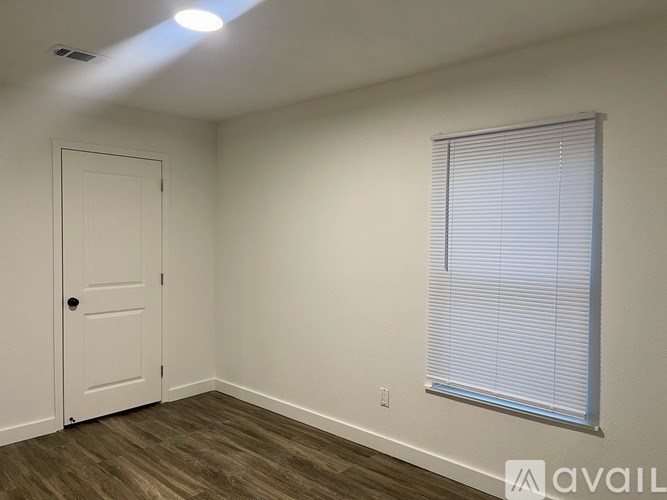 A room with a white door, a window with blinds, and a wooden floor.