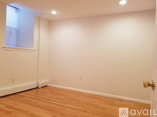 A room with wooden flooring and white walls, with a window on the left and a door on the right.
