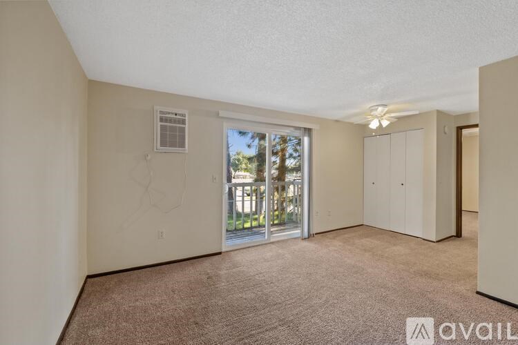 A room with beige walls and carpet, featuring a window with a view of trees and a building outside, and a sliding glass door leading to a balcony.