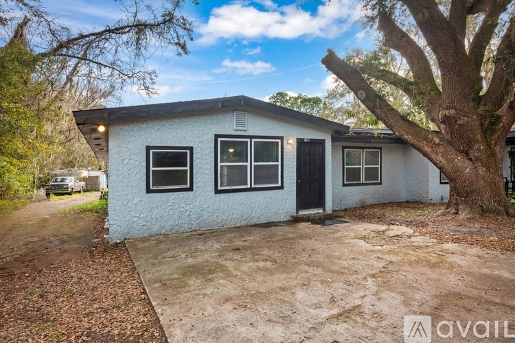 A white house with a black door and windows is surrounded by trees and has a gravel driveway.