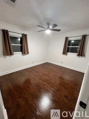 A room with brown wooden flooring and two windows with brown curtains.