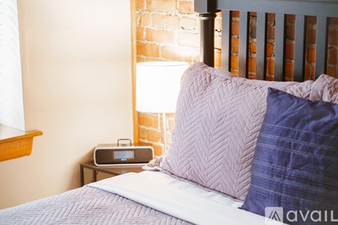 A bed with a white comforter and a purple pillow is in front of a brick wall.
