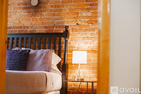 A bed with a white comforter and a blue pillow is in front of a brick wall with a lamp on the side.