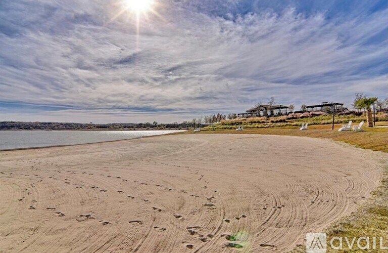 A beach with footprints in the sand leading to a row of trees and a building in the distance under a blue sky with the sun shining.
