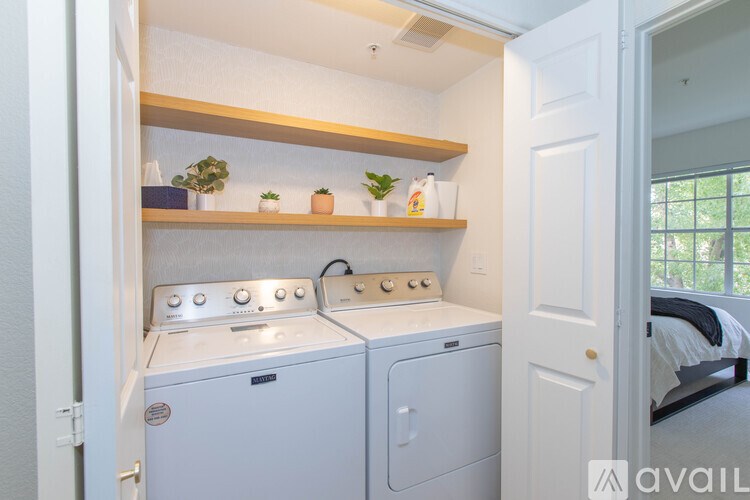A laundry room with a washer and dryer, a shelf with plants, and a doorway to a bedroom.