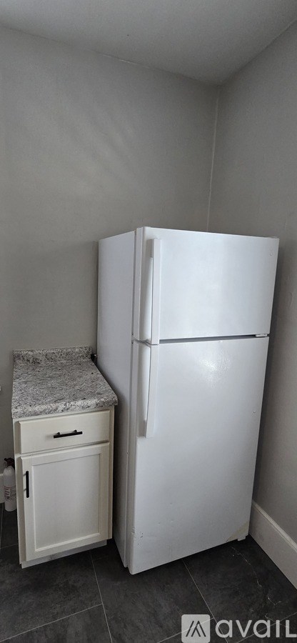 A white refrigerator stands next to a small white cabinet with a granite countertop.