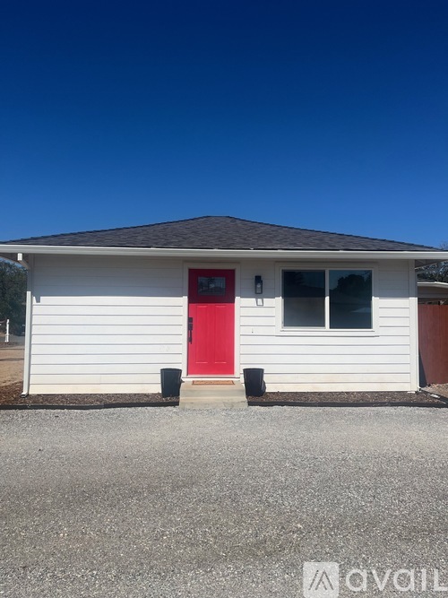 A small white house with a red door stands alone in a gravel lot.