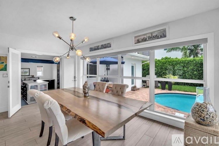 A dining room with a wooden table and chairs, a chandelier, and a view of a pool and greenery outside the window.