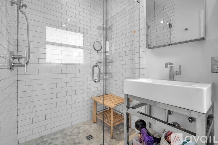 A bathroom with a white sink, a glass shower door, and a wooden stool.