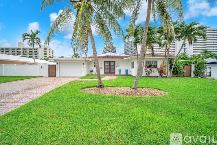A house with a white exterior and a brown roof is surrounded by a well-manicured lawn and palm trees.