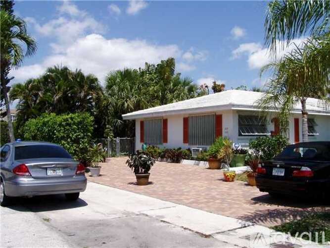 A house with a red and white exterior is surrounded by palm trees and has a car parked in front.