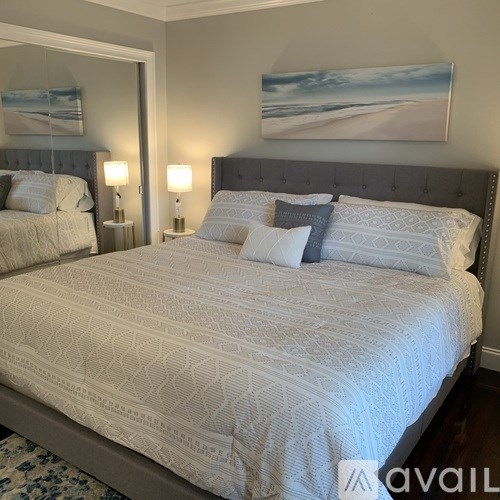 A bed with a white comforter and two blue pillows is in the foreground of a room with a painting on the wall.