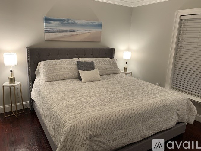 A bed with a white comforter and two pillows sits in a room with a painting on the wall and two lamps on either side of the bed.