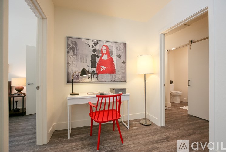 A red chair sits in front of a desk with a lamp and a picture on the wall.