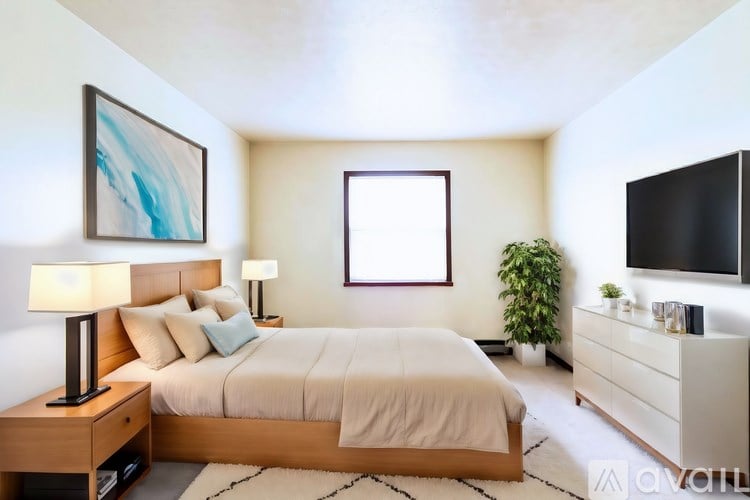 A well-lit bedroom with a large bed, a side table with a lamp, and a TV on the wall.