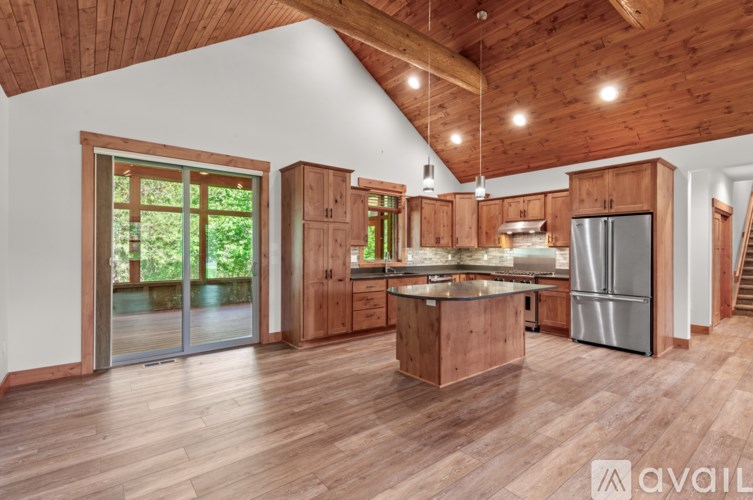 A kitchen with wooden floors and cabinets, a stainless steel refrigerator, and a large window with a view of greenery.