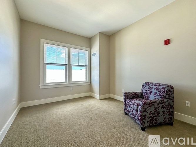 A room with a carpeted floor, a window with blinds, a wall-mounted fire extinguisher, and a chair with a floral pattern.