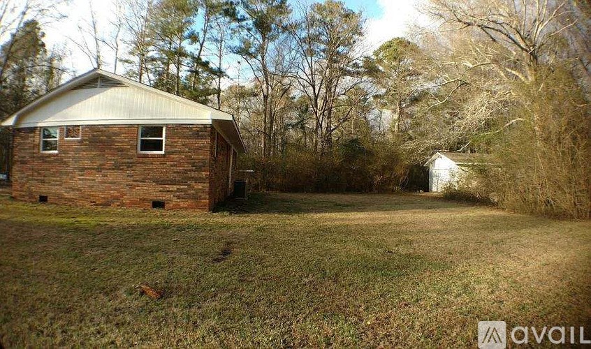 A small brick house with a metal roof sits in a grassy field with trees in the background.