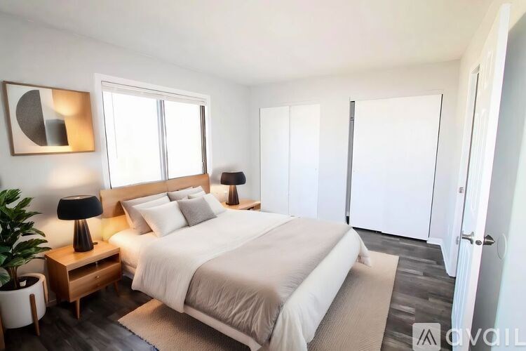 A bedroom with a large bed, a nightstand with a lamp, and a plant.