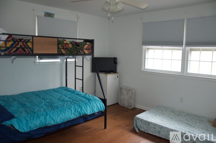 A bedroom with a blue bedspread on a bed, a bunk bed with a quilt, a window with blinds, a ceiling fan, a washing machine, and a wall-mounted TV.