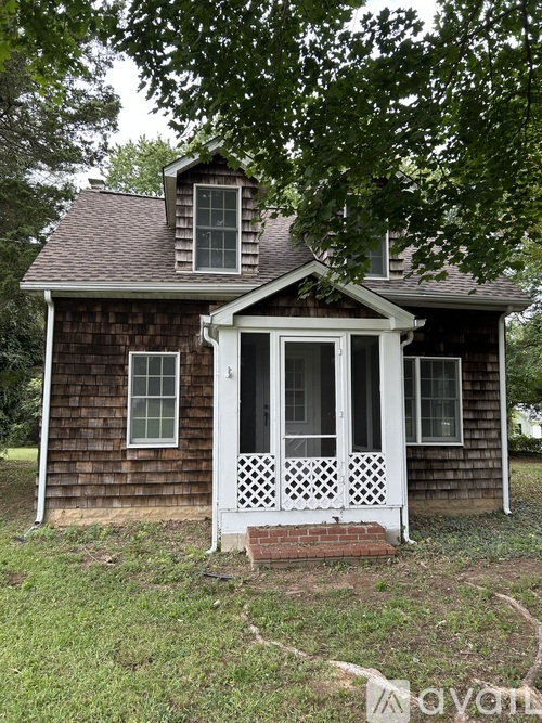 A small house with a porch and a brick base is surrounded by greenery.