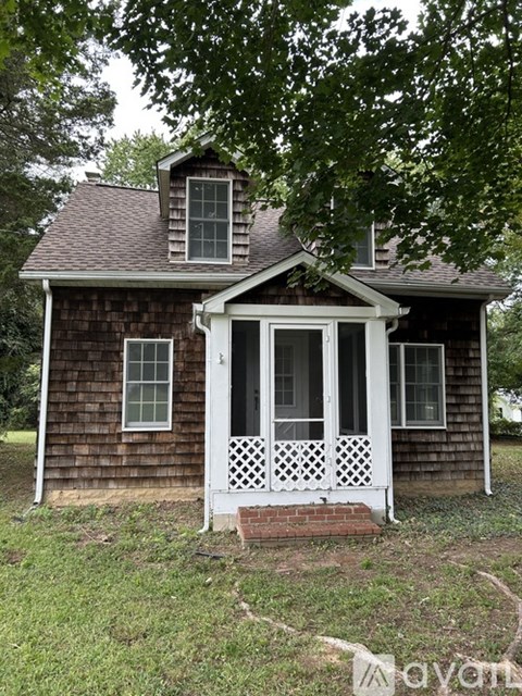 A small house with a porch and a brick base is surrounded by greenery.