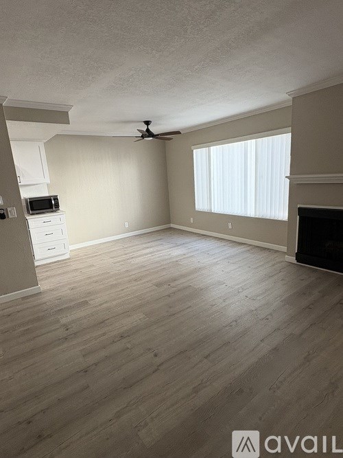 A spacious room with a ceiling fan and light, hardwood floors, and a window with blinds.