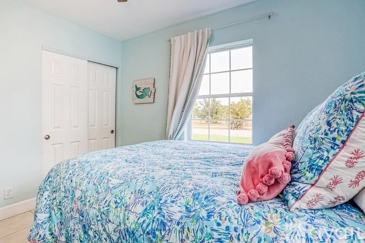 A bed with a blue and white comforter and a pink pillow is in the foreground of a room with light blue walls and a white door.