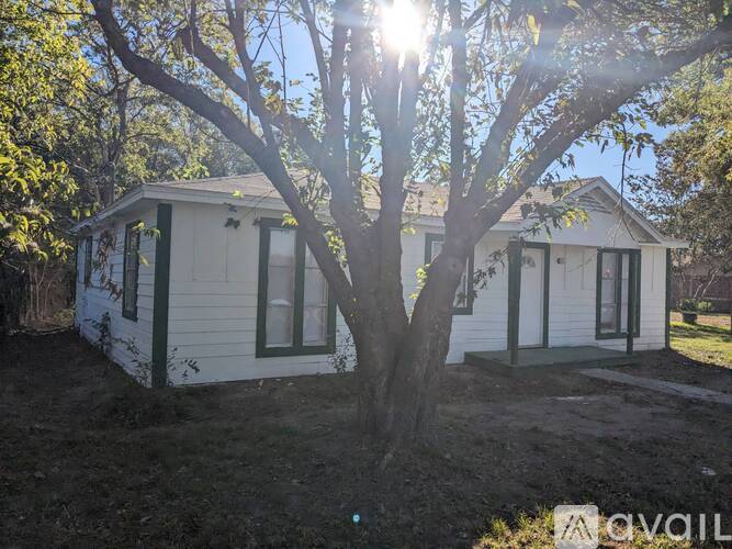 A white house with green trim is surrounded by trees and has a large tree in front of it.