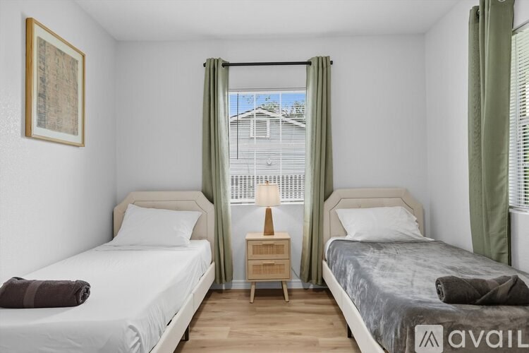 A bedroom with two beds, one with a white comforter and the other with a grey comforter, a nightstand with a lamp, and a window with green curtains.