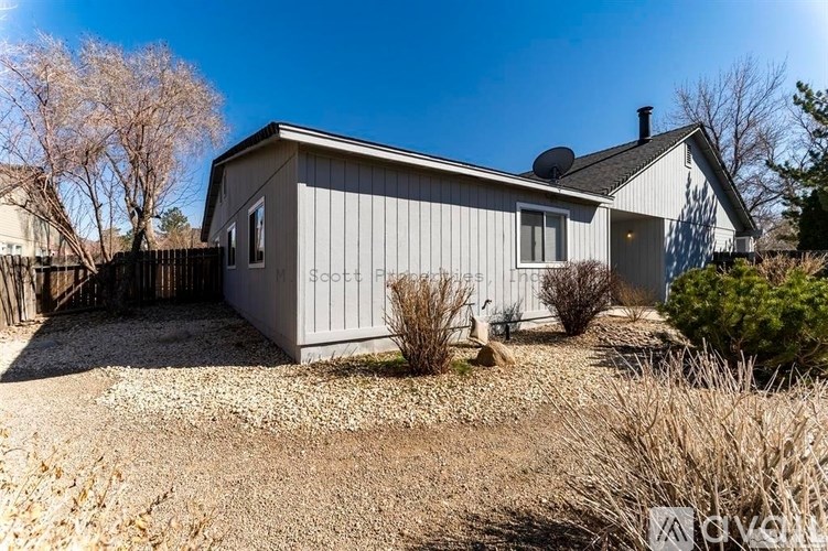 A house with a grey exterior and a brown roof is surrounded by a gravel driveway and some dry grass.