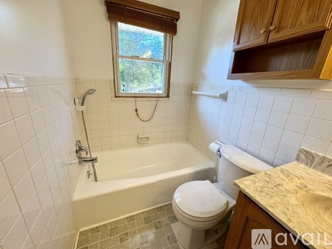 A white toilet sits in a bathroom with a white tub and a window.