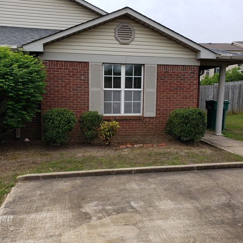 A small house with a window and a door surrounded by bushes and a driveway.