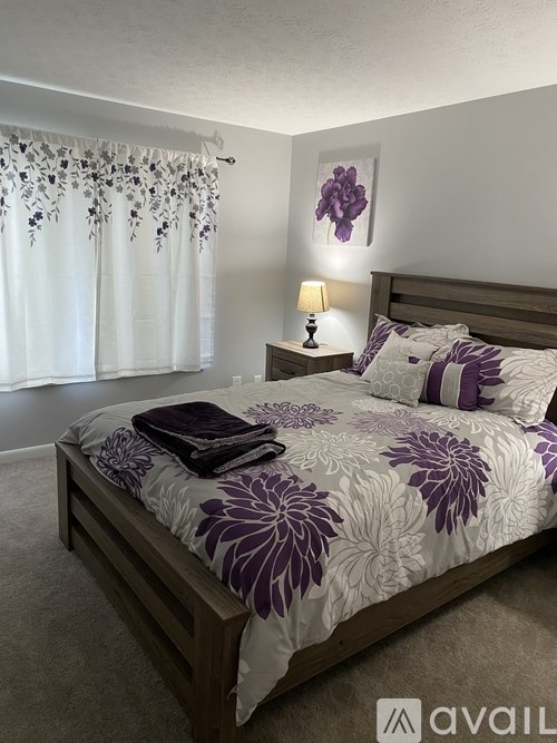 A bed with a purple and white comforter and matching pillows sits in a room with a white curtain and a lamp on a nightstand.