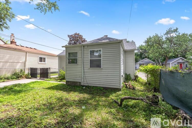 A small house with a grey siding is surrounded by greenery and other houses.