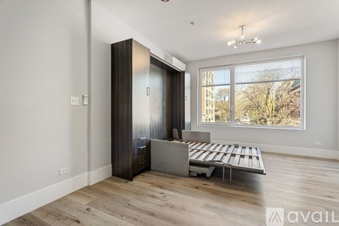 A room with a wooden floor, a white wall, a brown wardrobe, a bed with a grey headboard, and a window with a view of trees and buildings outside.