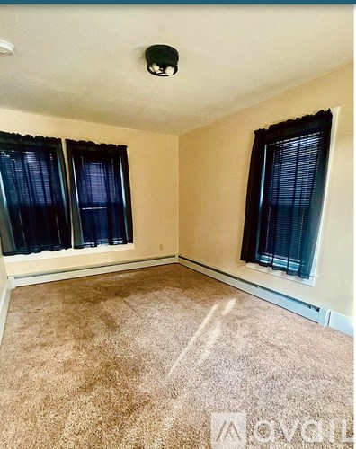 A room with beige walls, carpeted floor, and two windows with black curtains.