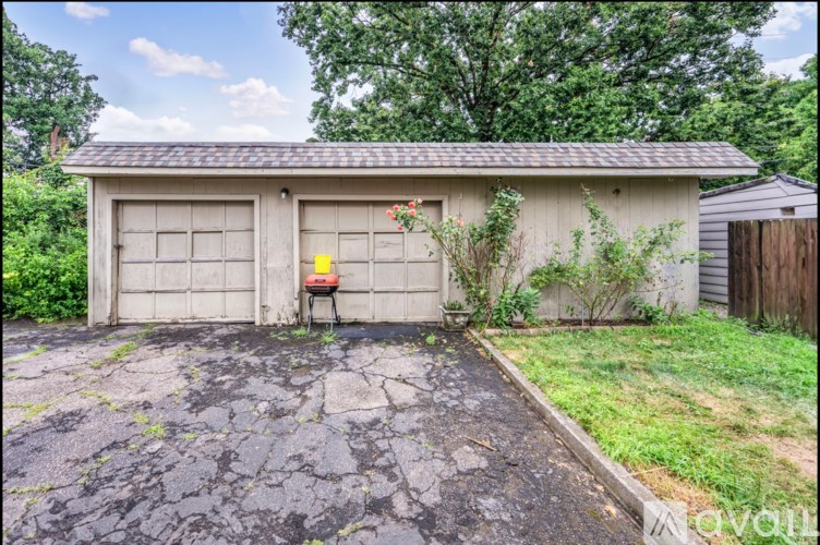 A small garage with a grey roof and a grey door is surrounded by a cracked driveway and green grass.